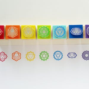 May include: A set of seven square rubber stamps with wooden bases, each featuring a unique geometric chakra symbol in red, orange, yellow, green, light blue, dark blue, and purple. Below the stamps are corresponding ink impressions on white paper.