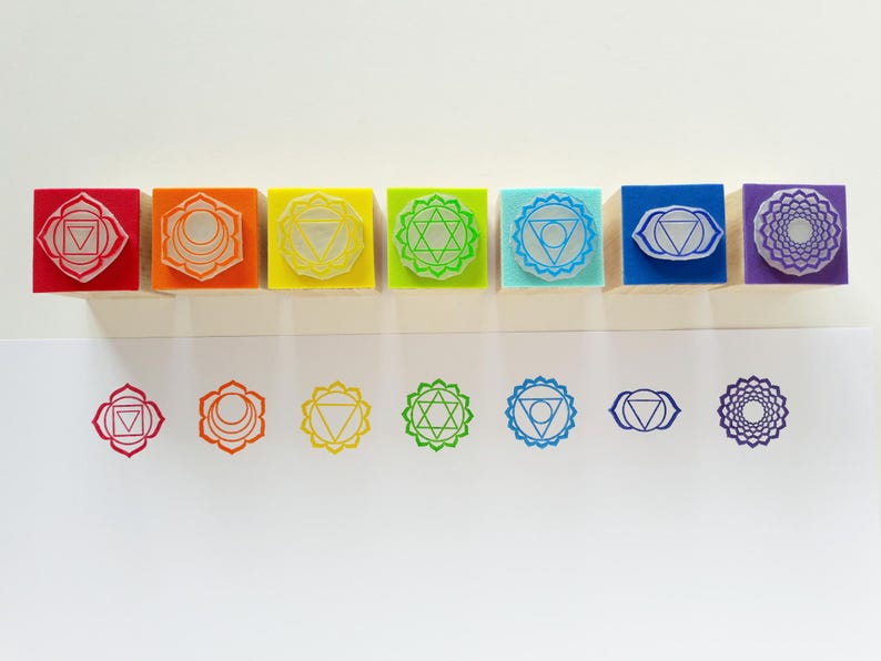 May include: A set of seven square rubber stamps with wooden bases, each featuring a unique geometric chakra symbol in red, orange, yellow, green, light blue, dark blue, and purple. Below the stamps are corresponding ink impressions on white paper.
