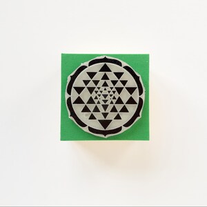 May include: A black and white rubber stamp featuring a Sri Yantra design, a geometric symbol used in Hinduism and Buddhism. The stamp is on a square of green foam, set against a white background.