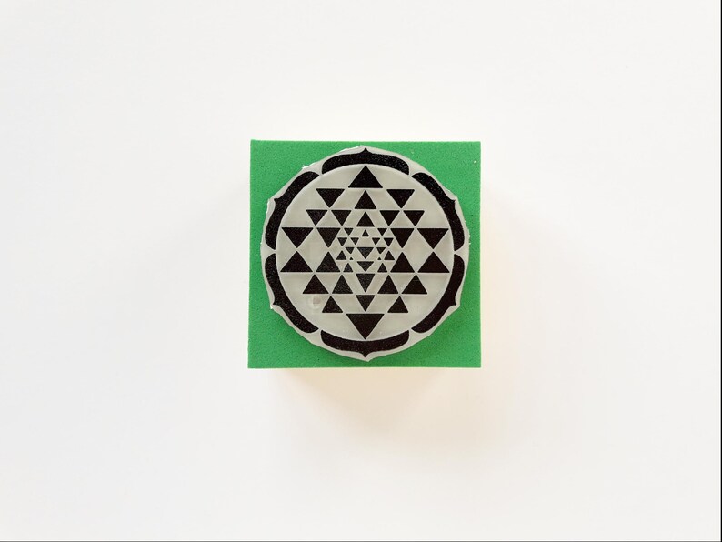 May include: A black and white rubber stamp featuring a Sri Yantra design, a geometric symbol used in Hinduism and Buddhism. The stamp is on a square of green foam, set against a white background.