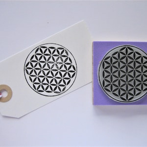 Op de afbeelding: Een paarse rubberen stempel met een zwart-wit geometrisch ontwerp van een levensbloem. De stempel staat op een witte achtergrond.