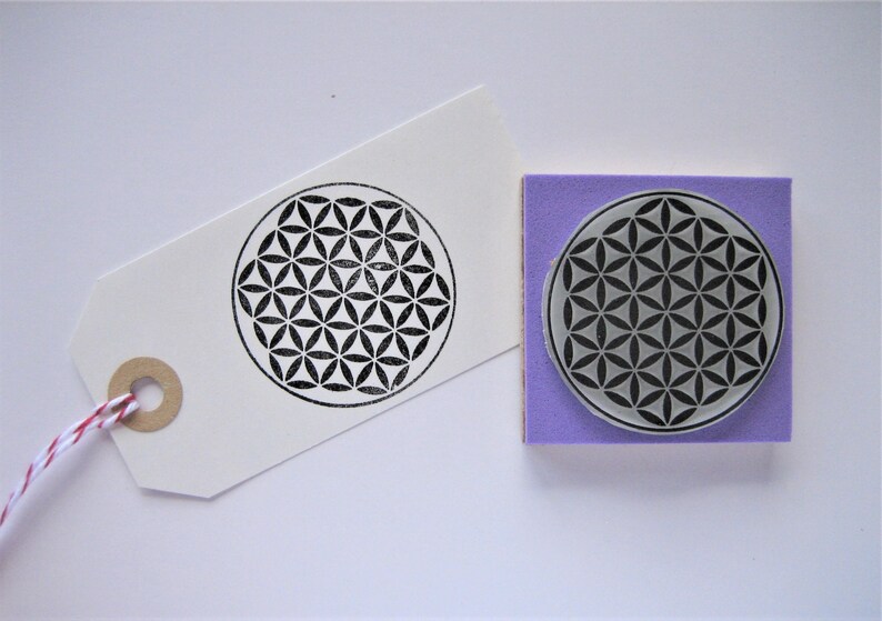 Op de afbeelding: Een paarse rubberen stempel met een zwart-wit geometrisch ontwerp van een levensbloem. De stempel staat op een witte achtergrond.