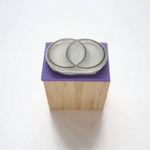 Op de afbeelding: Een houten blok met een paarse vilt bovenkant en een zilveren rubberen stempel met twee overlappende cirkels.