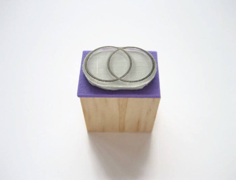 Op de afbeelding: Een houten blok met een paarse vilt bovenkant en een zilveren rubberen stempel met twee overlappende cirkels.