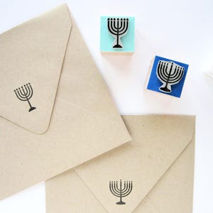 May include: Two light brown paper envelopes stamped with a black menorah design, accompanied by two square rubber stamps featuring the same menorah motif on light blue and dark blue wooden blocks, and a black ink pad.