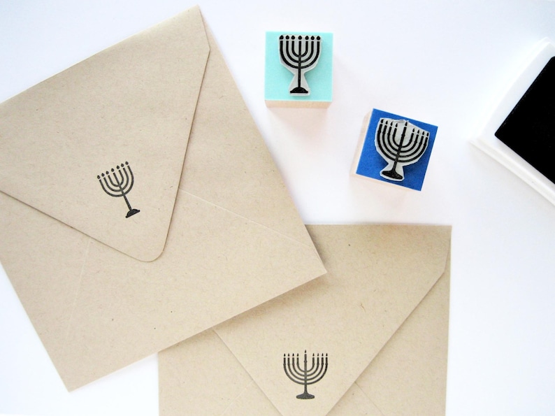 May include: Two light brown paper envelopes stamped with a black menorah design, accompanied by two square rubber stamps featuring the same menorah motif on light blue and dark blue wooden blocks, and a black ink pad.