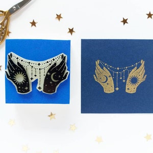 May include: Two hands holding a celestial necklace with a sun and moon charm. The hands are black and the necklace is white with gold stars. The image is on a blue background.
