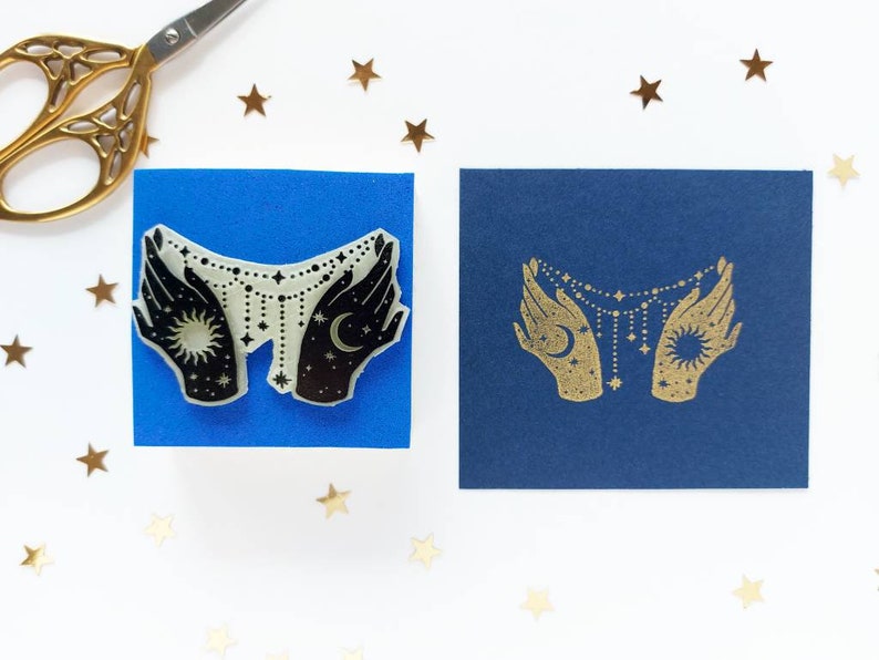 May include: Two hands holding a celestial necklace with a sun and moon charm. The hands are black and the necklace is white with gold stars. The image is on a blue background.