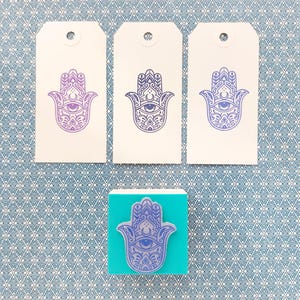 May include: Three rectangular white paper tags with a purple Hamsa hand print and a square teal rubber stamp featuring the same purple Hamsa hand design. Each tag has a circular hole at the top center for hanging or attachment purposes.