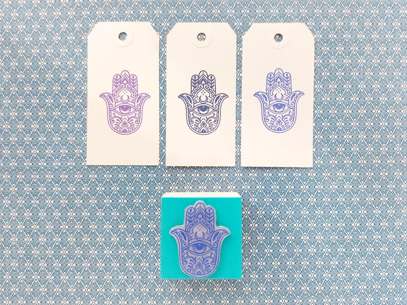 May include: Three rectangular white paper tags with a purple Hamsa hand print and a square teal rubber stamp featuring the same purple Hamsa hand design. Each tag has a circular hole at the top center for hanging or attachment purposes.