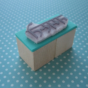 May include: A rectangular rubber stamp with a light wood base and a teal foam layer. The stamp features a raised, light purple rubber design with the text "DREAM" printed in a stylized, angular font on the top surface of the foam layer.