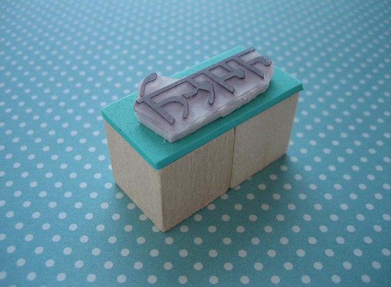 May include: A rectangular rubber stamp with a light wood base and a teal foam layer. The stamp features a raised, light purple rubber design with the text "DREAM" printed in a stylized, angular font on the top surface of the foam layer.