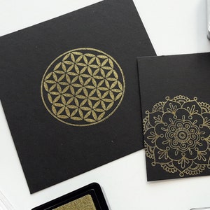Op de afbeelding: Twee zwarte kaarten met gouden geometrische ontwerpen. De eerste kaart heeft een levensbloemontwerp. De tweede kaart heeft een mandala-ontwerp.