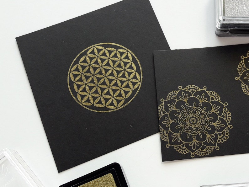 Op de afbeelding: Twee zwarte kaarten met gouden geometrische ontwerpen. De eerste kaart heeft een levensbloemontwerp. De tweede kaart heeft een mandala-ontwerp.