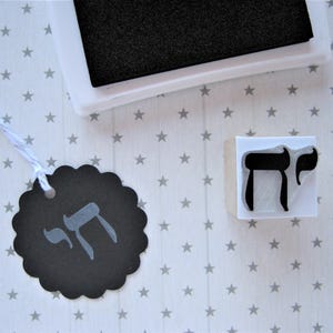 May include: A square rubber stamp featuring the Hebrew letter Chai in black ink, positioned next to a black scalloped paper tag with a white string. The tag displays a stamped impression of the same Hebrew character in a light gray color.
