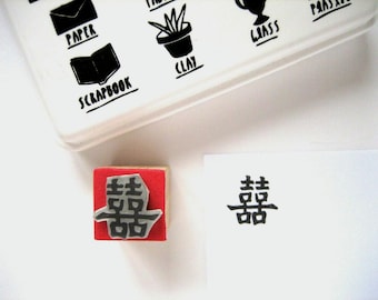 May include: A square rubber stamp with a red base and a wooden block, featuring the Chinese double happiness character. Next to it is a white paper with the same character stamped in black ink. Above, a white container shows icons for paper, clay, and scrapbook.