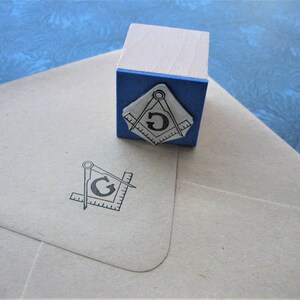 May include: A wooden stamp with a blue base and a Masonic symbol. The stamp is next to a light-colored envelope with the same symbol printed on it. The symbol features a compass, a square, and the letter "G".