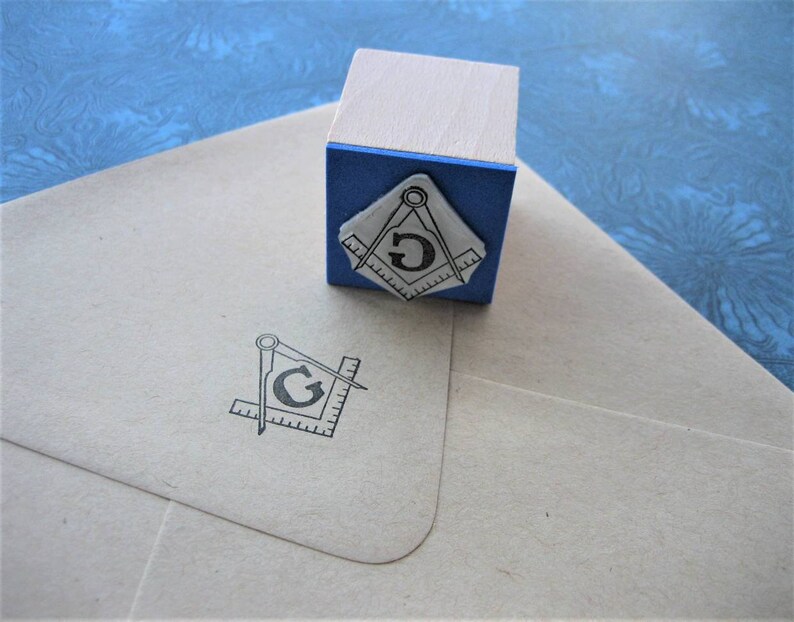 May include: A wooden stamp with a blue base and a Masonic symbol. The stamp is next to a light-colored envelope with the same symbol printed on it. The symbol features a compass, a square, and the letter "G".