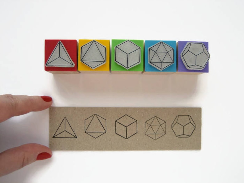 May include: Six colorful wooden blocks with different geometric shapes printed on them. The shapes include a triangle, a cube, and a dodecahedron. The blocks are arranged in a row from left to right. Below the blocks is a brown card with the same geometric shapes printed on it.