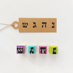May include: A rectangular brown paper tag with a string loop, printed with four black Hebrew letters. Below the tag are four square wooden stamps with colorful foam bases in purple, blue, pink, and green, each featuring a raised Hebrew letter.