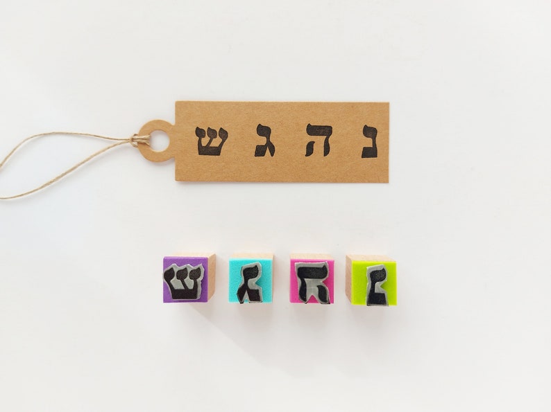 May include: A rectangular brown paper tag with a string loop, printed with four black Hebrew letters. Below the tag are four square wooden stamps with colorful foam bases in purple, blue, pink, and green, each featuring a raised Hebrew letter.