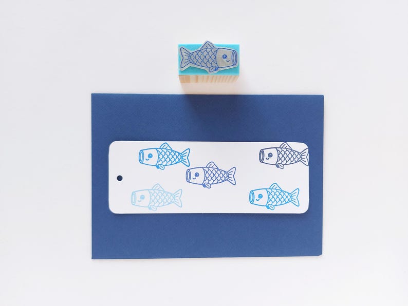 May include: A blue fish stamp with a wooden handle sits above a navy blue card with a white tag. The tag features multiple blue fish illustrations. The stamp and illustrations are in a cartoon style.