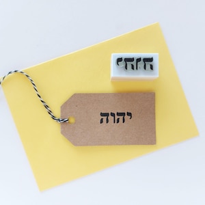 May include: A rectangular rubber stamp with a white base and black Hebrew text, positioned next to a brown paper gift tag featuring the same Hebrew text in black ink. The tag is attached to a black and white twisted twine string.