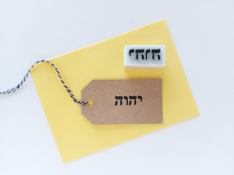 May include: A rectangular rubber stamp with a white base and black Hebrew text, positioned next to a brown paper gift tag featuring the same Hebrew text in black ink. The tag is attached to a black and white twisted twine string.