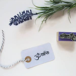 Puede incluir: Un sello de goma morado con el texto "Om Shanti" en s&aacute;nscrito y un peque&ntilde;o s&iacute;mbolo Om. El sello est&aacute; sobre una superficie blanca con una cuerda blanca y negra y una rama de lavanda.