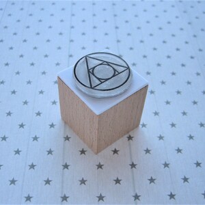 May include: A wooden block with a white paper surface. The paper surface has a clear, round rubber stamp with a black geometric design. The design is a triangle with a circle inside.