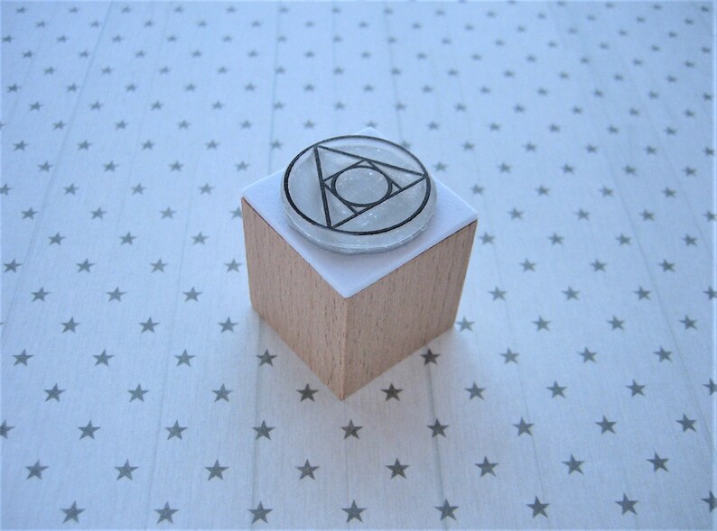 May include: A wooden block with a white paper surface. The paper surface has a clear, round rubber stamp with a black geometric design. The design is a triangle with a circle inside.