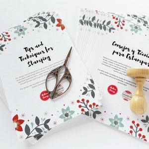 May include: A stack of white cards with a red and green floral pattern. The cards have the text "Tips and Techniques for Stamping" and "Consejos y T&eacute;cnicas para Estampar" printed on them. A pair of scissors and a wooden stamp are also in the image.