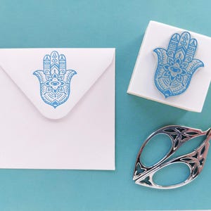 May include: A white square envelope and a square rubber stamp, both featuring a blue Hamsa hand design with intricate patterns. A pair of silver-toned metal scissors with decorative cutouts in the handles is positioned nearby.