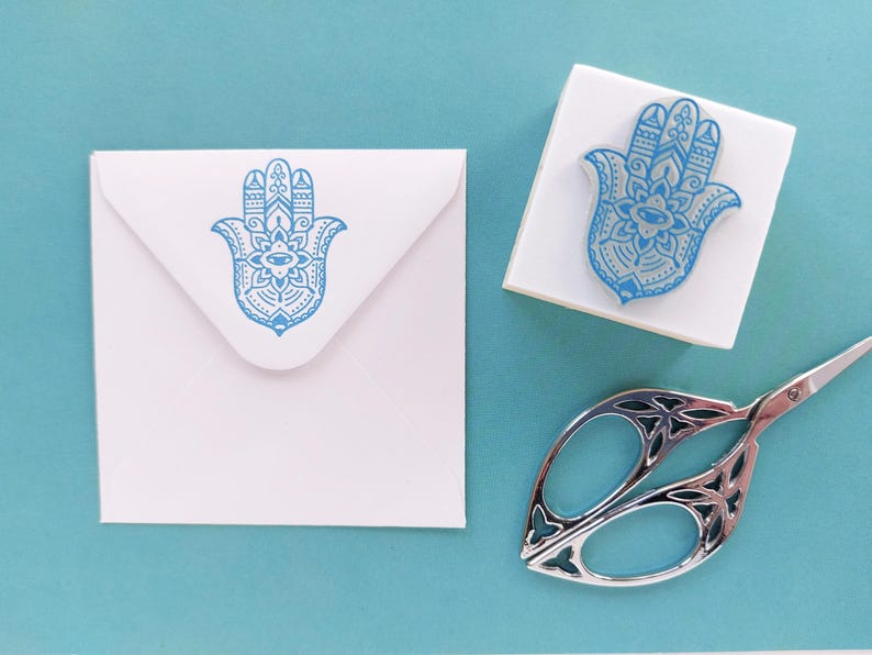 May include: A white square envelope and a square rubber stamp, both featuring a blue Hamsa hand design with intricate patterns. A pair of silver-toned metal scissors with decorative cutouts in the handles is positioned nearby.