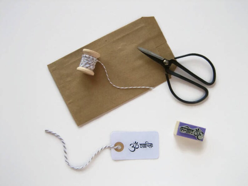 Puede incluir: Una bolsa de papel marr&oacute;n, un carrete de hilo blanco y negro, unas tijeras de podar negras, una etiqueta blanca con el texto "ॐ शांति" en negro y un sello de goma morado con el texto "ॐ शांति" en negro.