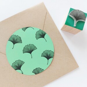 May include: A craft image featuring a round, light green sticker with a black ginkgo leaf pattern, placed on a brown envelope. A wooden stamp with a ginkgo leaf design sits nearby. The stamp has a green base.