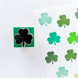 May include: A green rubber stamp with a black shamrock design. The stamp is on a white surface next to a sheet of paper with multiple green shamrocks printed on it.
