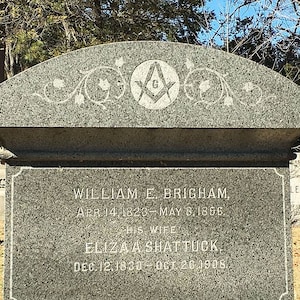 May include: A gray granite headstone with the inscription "WILLIAM E. BRIGHAM" and "ELIZA A SHATTUCK". The top of the headstone features a Masonic symbol and decorative floral designs. Dates of birth and death are also inscribed.