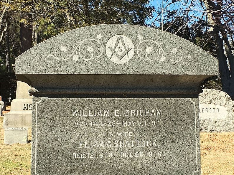 May include: A gray granite headstone with the inscription "WILLIAM E. BRIGHAM" and "ELIZA A SHATTUCK". The top of the headstone features a Masonic symbol and decorative floral designs. Dates of birth and death are also inscribed.