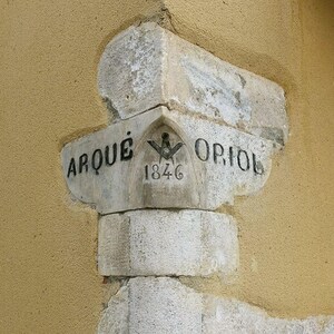 May include: A weathered stone corner piece with the words "ARQUE" and "ORIOL" inscribed on it. The year "1846" is visible, along with a symbol. The stone is a light gray color and is set against a textured, tan wall.