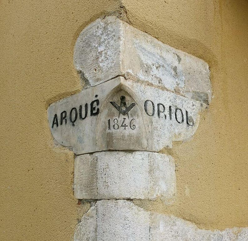 May include: A weathered stone corner piece with the words "ARQUE" and "ORIOL" inscribed on it. The year "1846" is visible, along with a symbol. The stone is a light gray color and is set against a textured, tan wall.