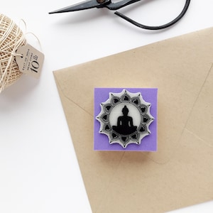 May include: A square purple rubber stamp featuring a black silhouette of a seated Buddha figure centered within a white lotus flower design. The stamp is positioned on a tan envelope next to a ball of light brown hemp cord and black scissors.