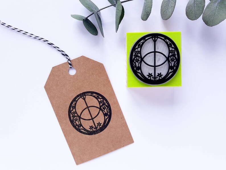 Op de afbeelding: Een zwarte en witte rubberen stempel met een cirkelvormig ontwerp met twee overlappende cirkels met een kruis erin. De stempel is gemonteerd op een felgroene basis. De stempel ligt op een wit oppervlak.