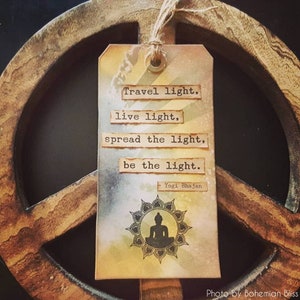 May include: A brown wooden peace sign with a beige tag hanging from it. The tag has a watercolour background and a black silhouette of a meditating Buddha. The tag has the text "Travel light, live light, spread the light, be the light. - Yogi Bhajan" printed on it.