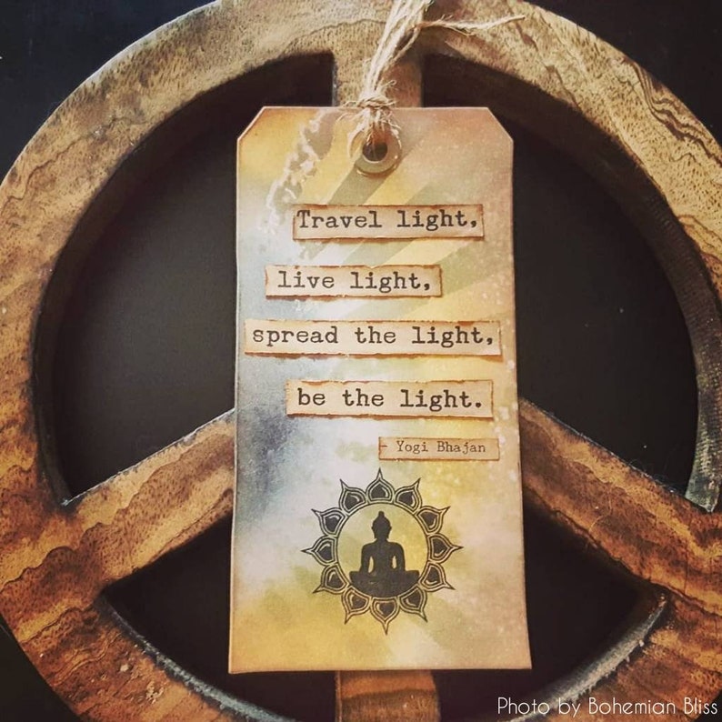 May include: A brown wooden peace sign with a beige tag hanging from it. The tag has a watercolour background and a black silhouette of a meditating Buddha. The tag has the text "Travel light, live light, spread the light, be the light. - Yogi Bhajan" printed on it.