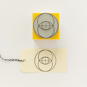 Op de afbeelding: Een gele houten blok met een zwart-wit geometrisch ontwerp van overlappende cirkels. Het ontwerp is ook afgedrukt op een cr&egrave;mekleurig label.