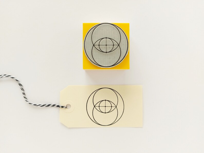 Op de afbeelding: Een gele houten blok met een zwart-wit geometrisch ontwerp van overlappende cirkels. Het ontwerp is ook afgedrukt op een cr&egrave;mekleurig label.