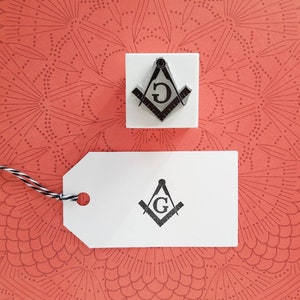 May include: A white tag with a black and white image of a compass and square with a "G" in the center. The tag is tied with a black and white string to a white square with a similar image of a compass and square with a "G" in the center.