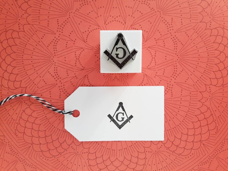 May include: A white tag with a black and white image of a compass and square with a "G" in the center. The tag is tied with a black and white string to a white square with a similar image of a compass and square with a "G" in the center.