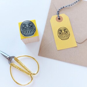 May include: A Daruma doll stamp on a yellow block, a matching tag, and gold scissors are arranged on a white surface. The stamp and tag feature a black Daruma doll design. A brown envelope is in the background.